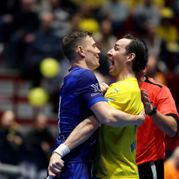 This image captures an intense moment during a floorball match, likely from the WFC 2024. It appears to show a heated exchange or confrontation between two players—one from each team—while a referee steps in to separate them. The player in blue and the player in yellow are face-to-face, with emotions clearly running high, as indicated by their expressions and body language.